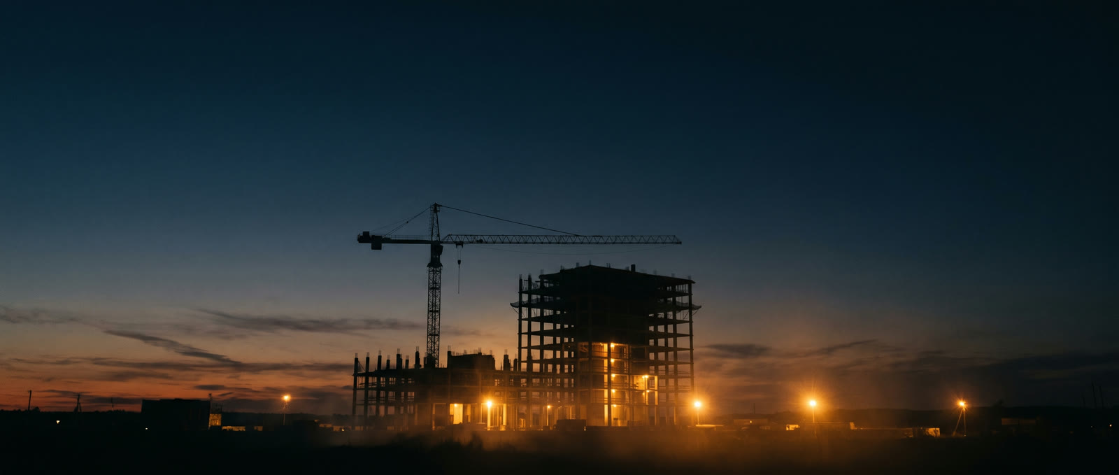 Silueta de un edificio en construcción con grúa al anochecer, cielo navy y luces cálidas de obra